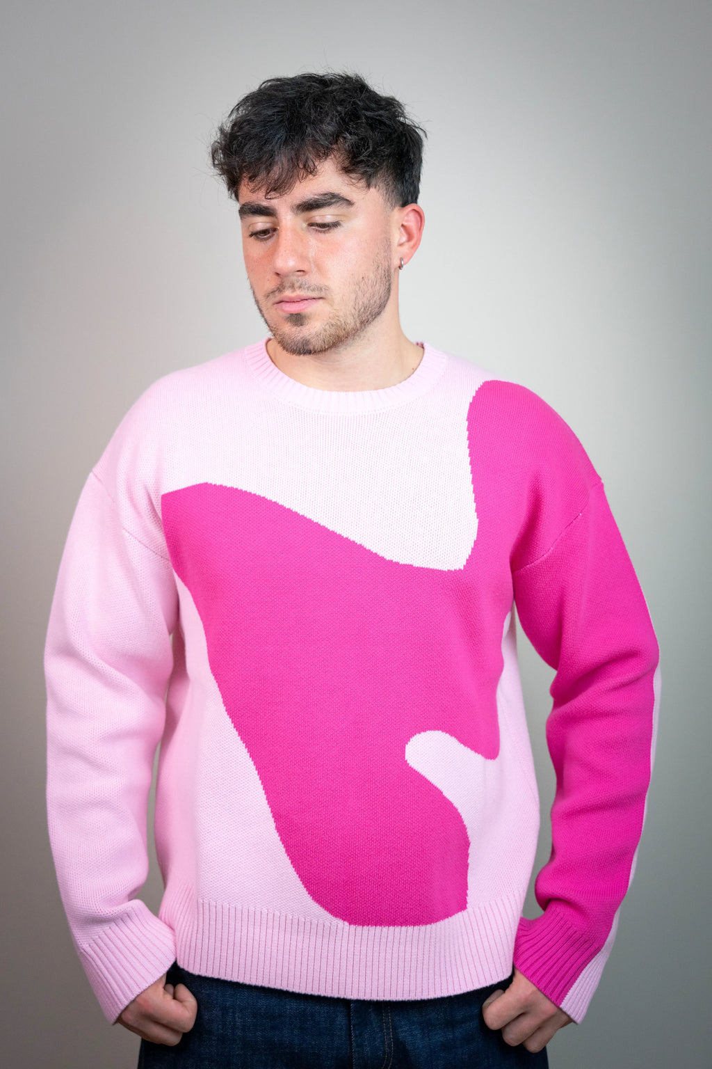 Hombre con suéter rosa claro y diseño abstracto fucsia, fondo gris neutro, moda casual