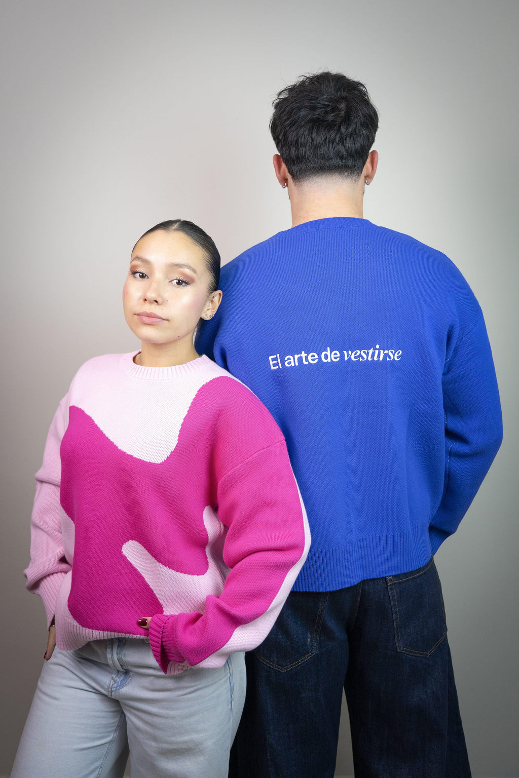 Mujer con suéter rosa y hombre con suéter azul que dice 'El arte de vestirse', moda casual.