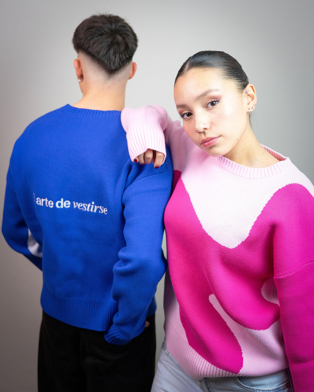 Pareja joven con suéteres modernos, ella en rosa y él en azul con texto 'arte de vestirse'.